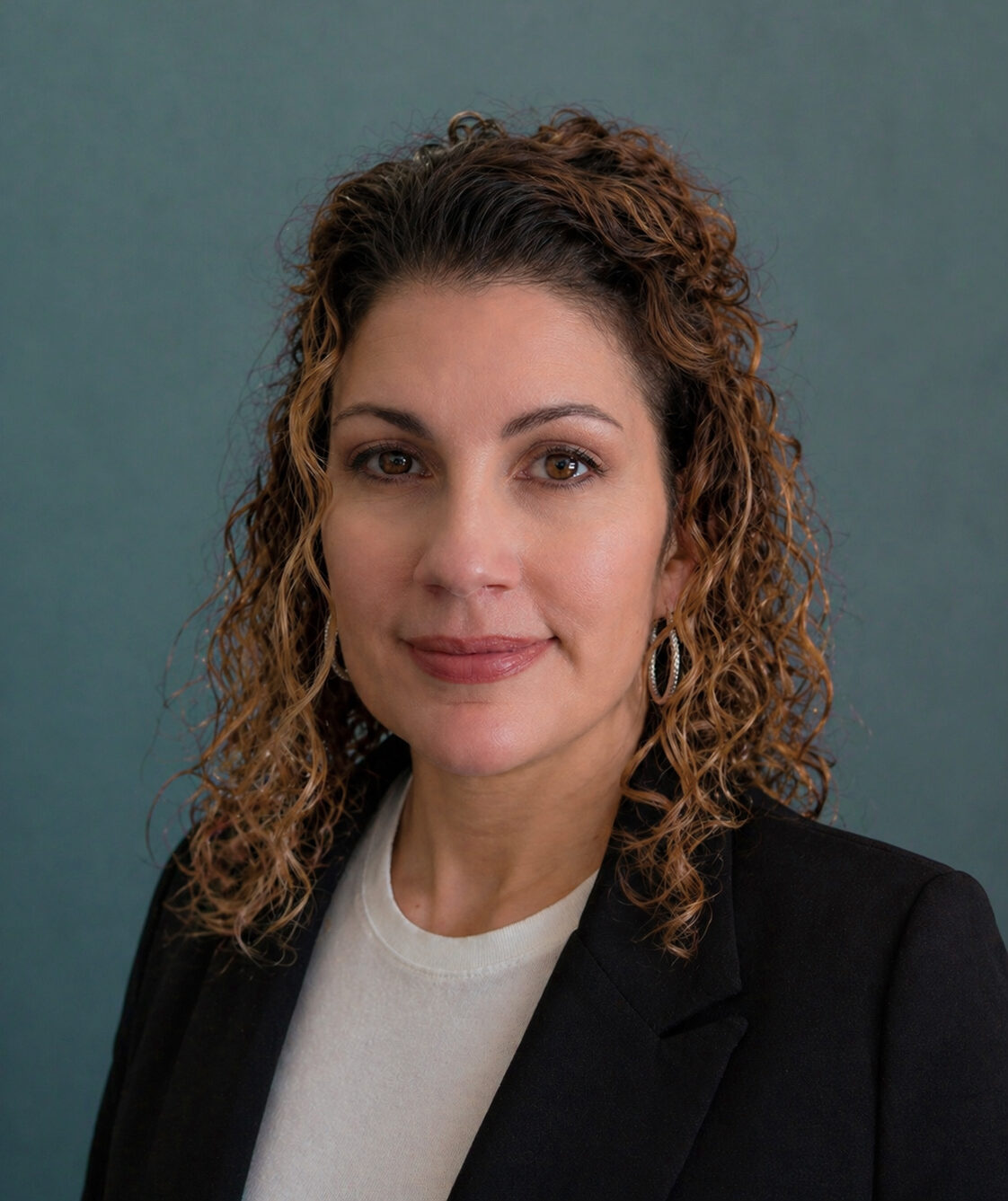 Head-and-shoulders portrait of a woman with curly brown hair, wearing a black blazer and white top, facing the camera.