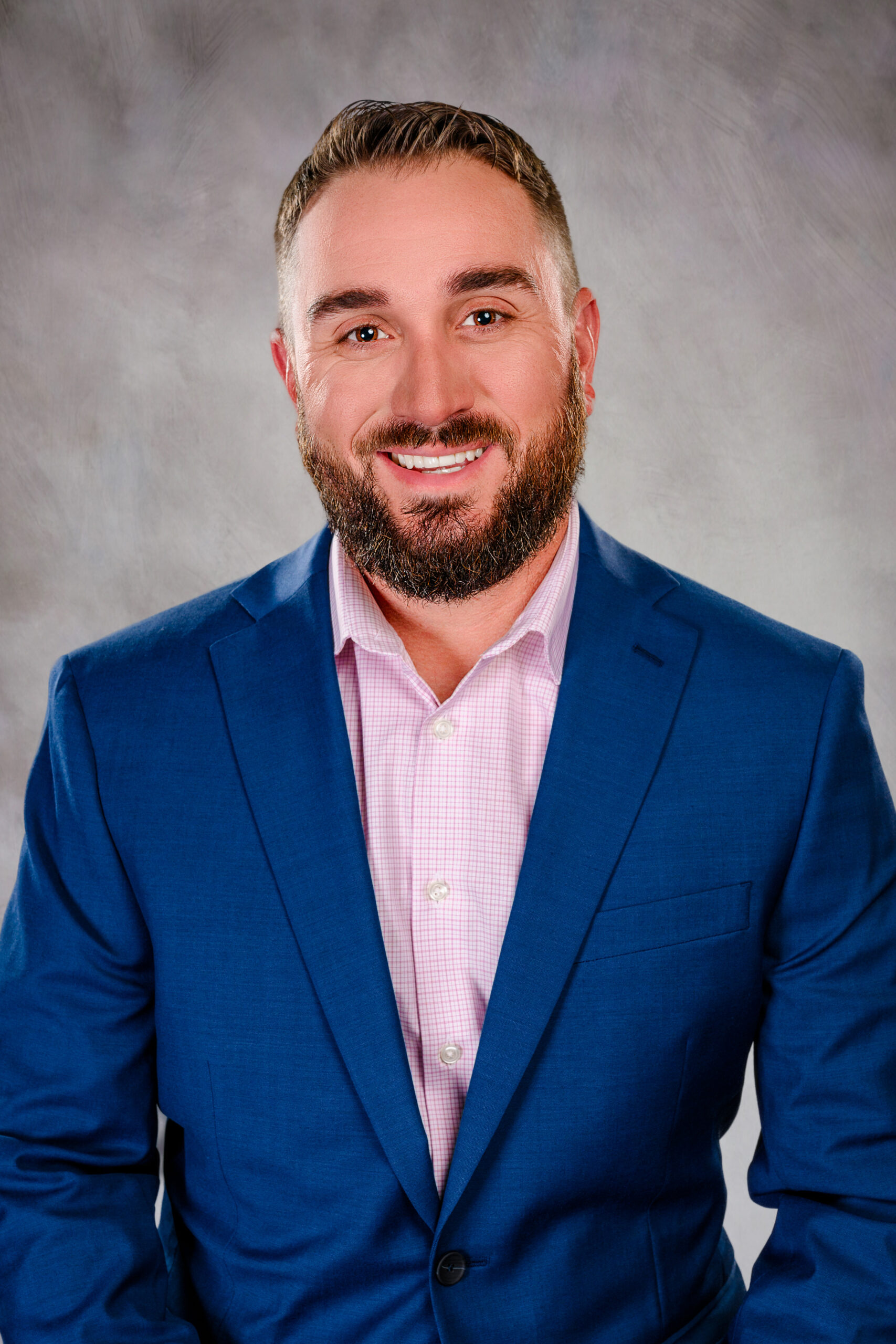 Smiling man with a beard in a blue blazer and pink check shirt, posed against a neutral gray backdrop.